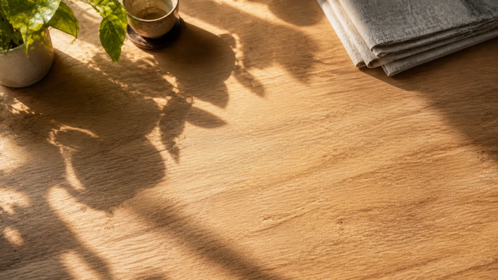 A warm timber table in side light, with a plant, a ceramic cup and a folded newspaper at its edges.